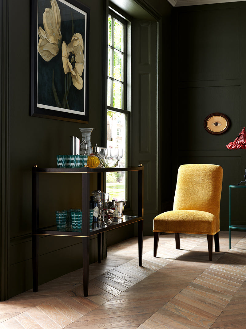 Nina Campbell Rita Slipper Chair, in a yellow velvet fabric, photographed in a room with dark green walls and wooden floors.